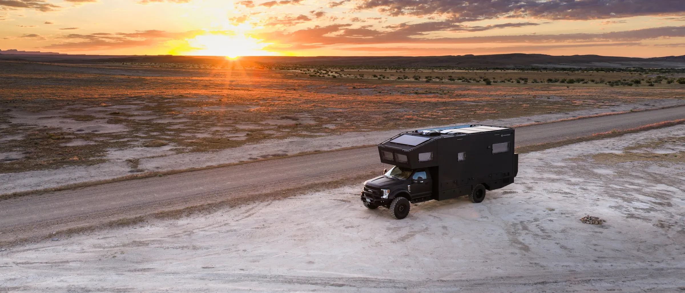 Aerial view of Emma's expedition RV crossing a desert plain at sunset